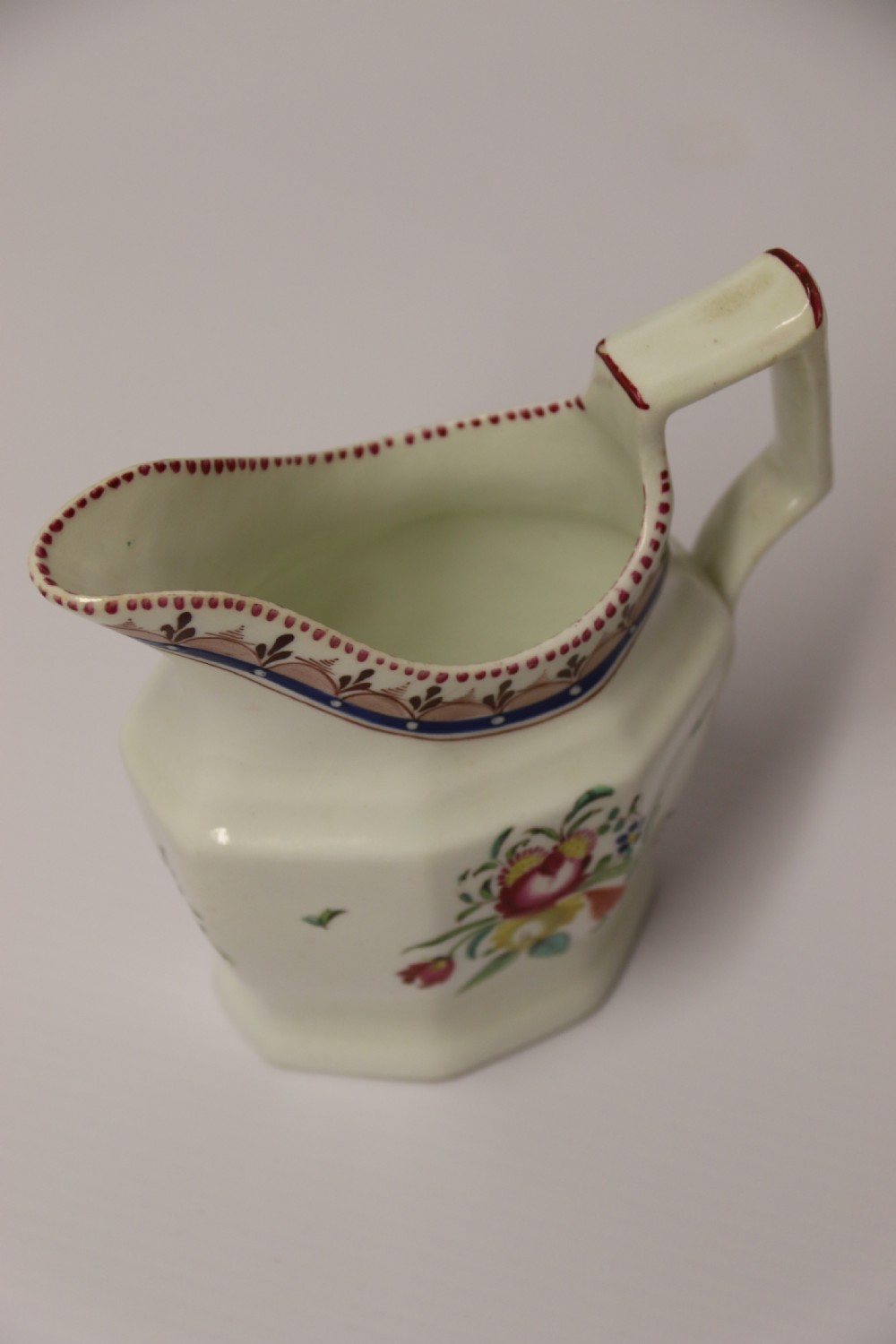 An 18th-century cream-coloured porcelain jug featuring hand-painted floral motifs, a decorated rim border, and an angular handle. View XIII