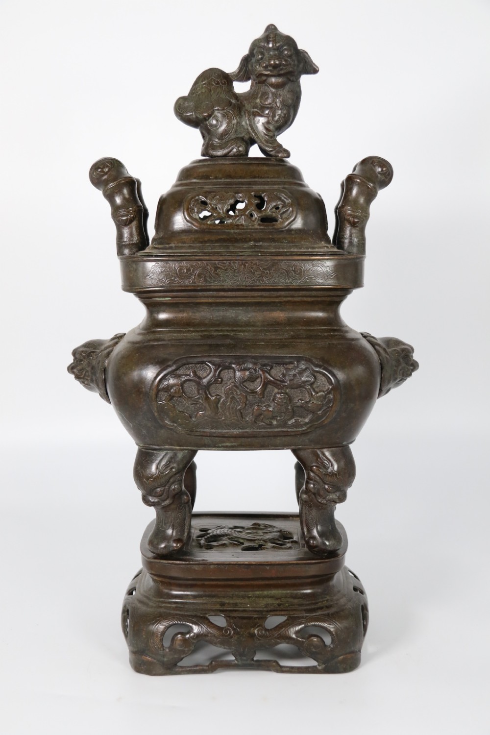 A 19th-century Chinese cast bronze incense censer featuring a lid surmounted by a guardian lion finial, relief panels, and animal-mask handles, set upon a matching bronze base.