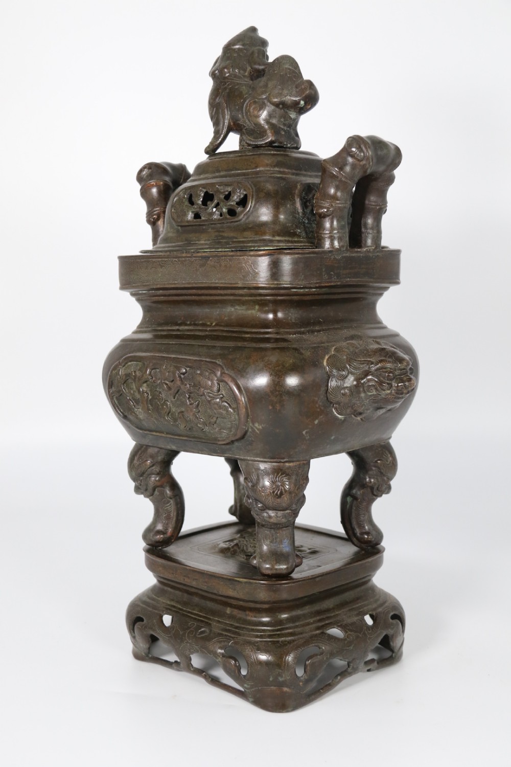 A 19th-century Chinese cast bronze incense censer featuring a lid surmounted by a guardian lion finial, relief panels, and animal-mask handles, set upon a matching bronze base. View IV