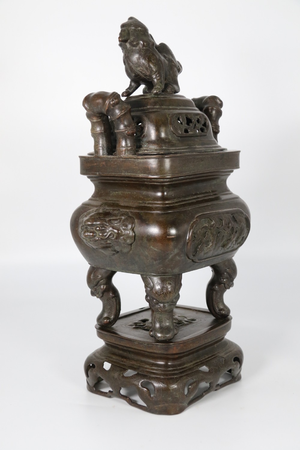 A 19th-century Chinese cast bronze incense censer featuring a lid surmounted by a guardian lion finial, relief panels, and animal-mask handles, set upon a matching bronze base. View V