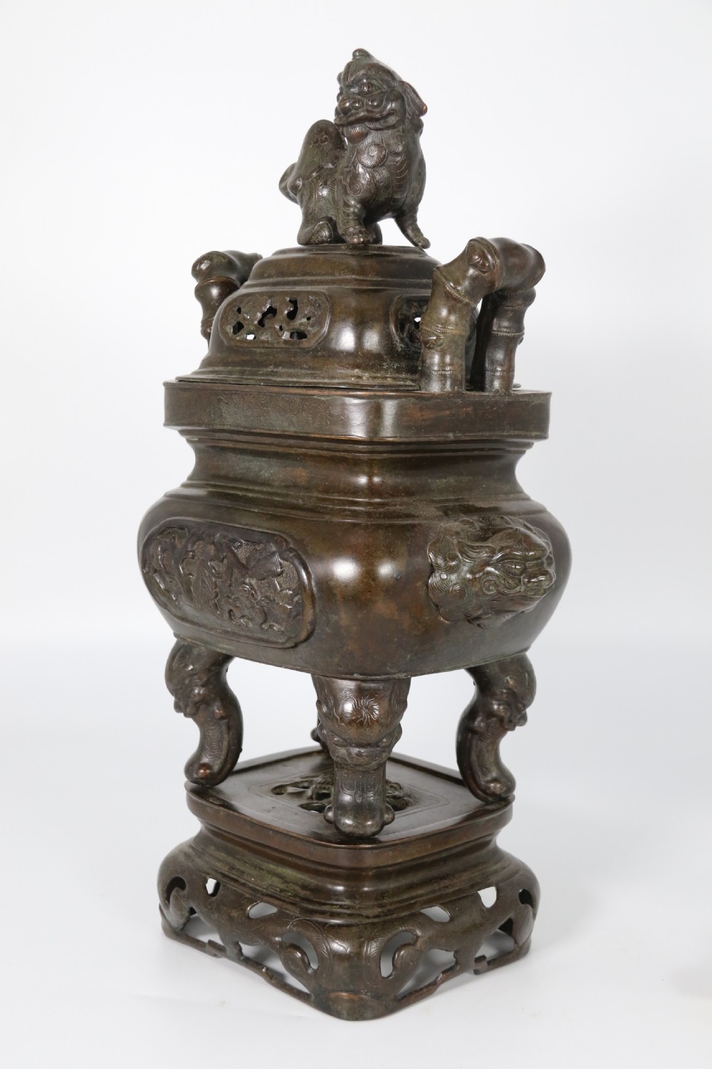 A 19th-century Chinese cast bronze incense censer featuring a lid surmounted by a guardian lion finial, relief panels, and animal-mask handles, set upon a matching bronze base. View VIII