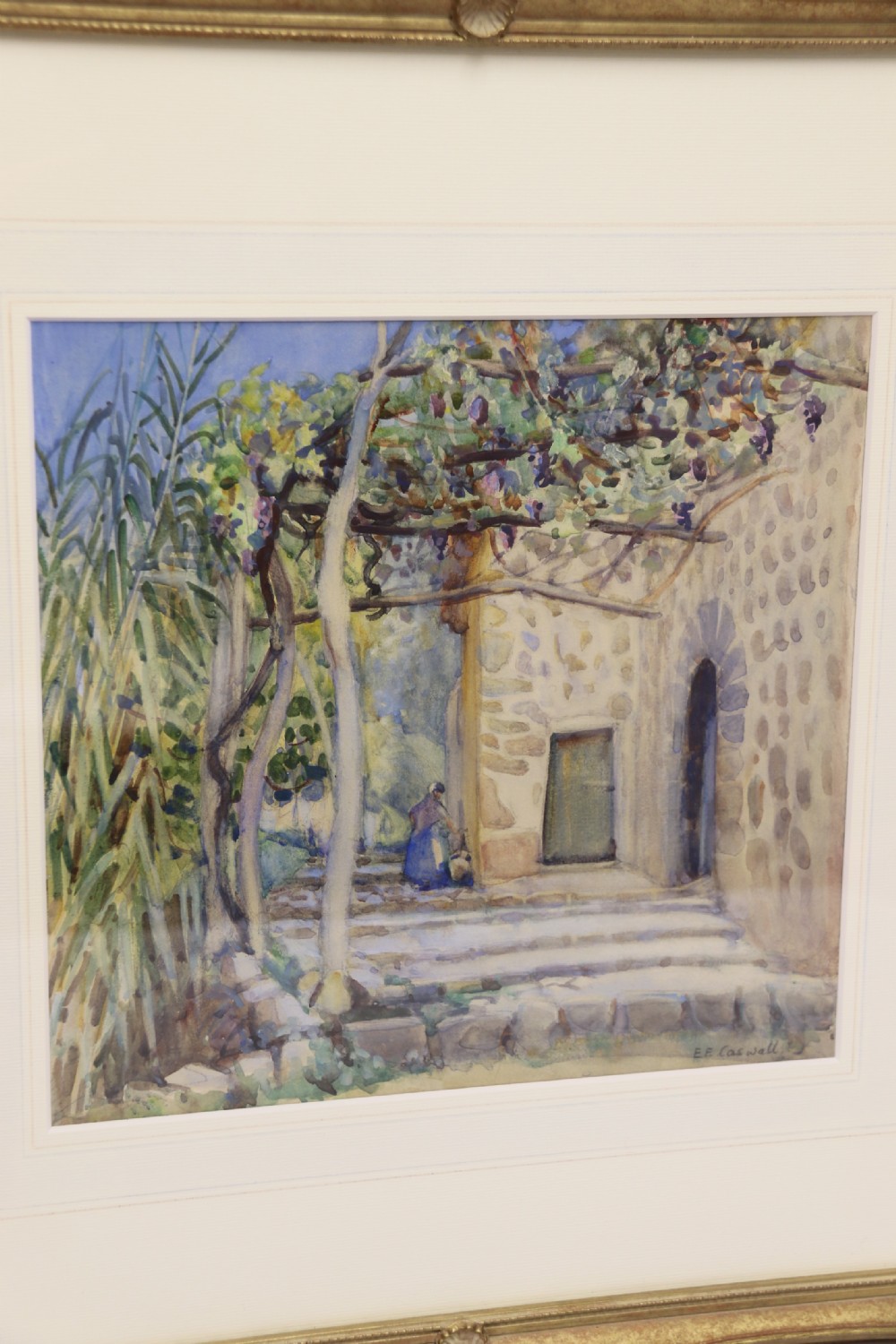 A framed 1920s watercolour landscape painting showing a stone building with vine foliage, set inside a decorative gilt frame with cream matting. View II