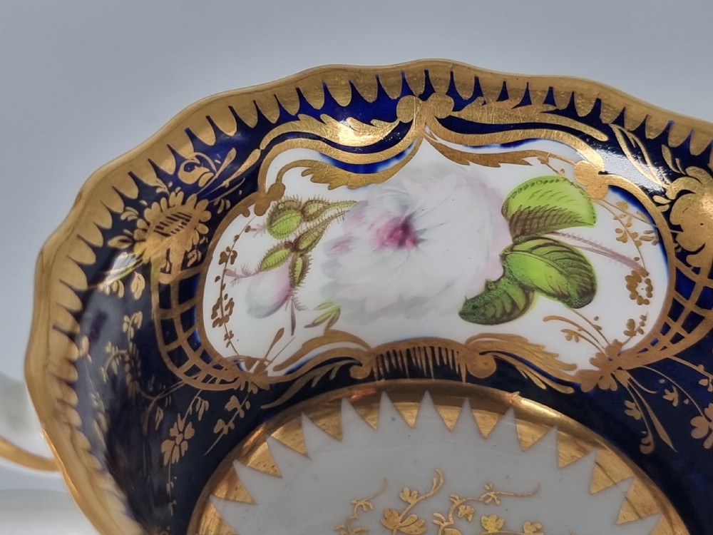 An early nineteenth-century Coalport porcelain cabinet trio featuring a teacup, coffee cup, and saucer styled with cobalt blue borders, gilded detailing, and hand-painted white rose panels. View XI