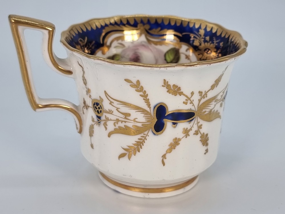 An early nineteenth-century Coalport porcelain cabinet trio featuring a teacup, coffee cup, and saucer styled with cobalt blue borders, gilded detailing, and hand-painted white rose panels. View XVI
