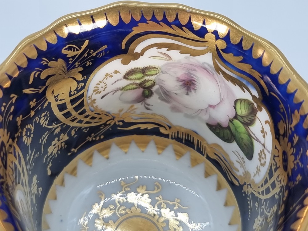 An early nineteenth-century Coalport porcelain cabinet trio featuring a teacup, coffee cup, and saucer styled with cobalt blue borders, gilded detailing, and hand-painted white rose panels. View XX