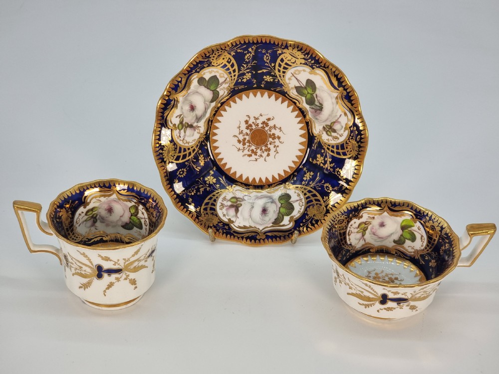 An early nineteenth-century Coalport porcelain cabinet trio featuring a teacup, coffee cup, and saucer styled with cobalt blue borders, gilded detailing, and hand-painted white rose panels. View XXIV