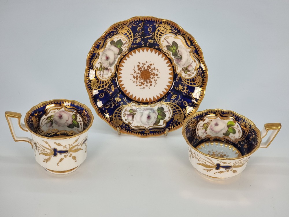 An early nineteenth-century Coalport porcelain cabinet trio featuring a teacup, coffee cup, and saucer styled with cobalt blue borders, gilded detailing, and hand-painted white rose panels. View XXV