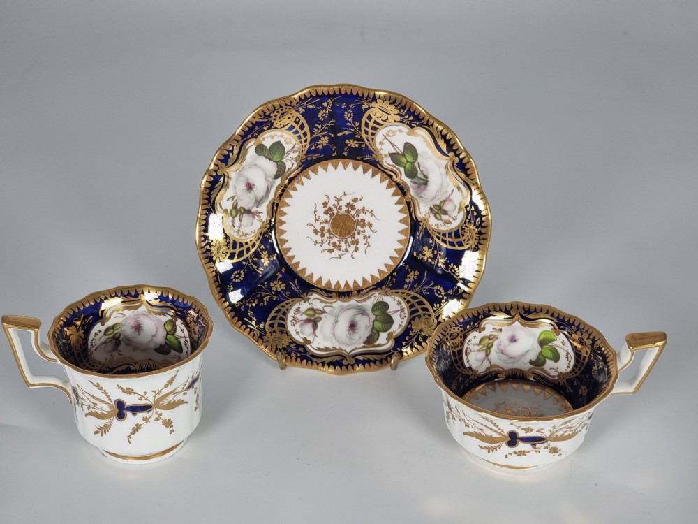 An early nineteenth-century Coalport porcelain cabinet trio featuring a teacup, coffee cup, and saucer styled with cobalt blue borders, gilded detailing, and hand-painted white rose panels. View XXVI