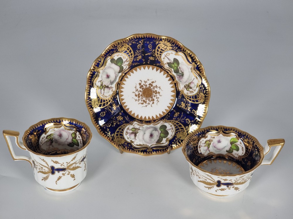 An early nineteenth-century Coalport porcelain cabinet trio featuring a teacup, coffee cup, and saucer styled with cobalt blue borders, gilded detailing, and hand-painted white rose panels. View XXVII