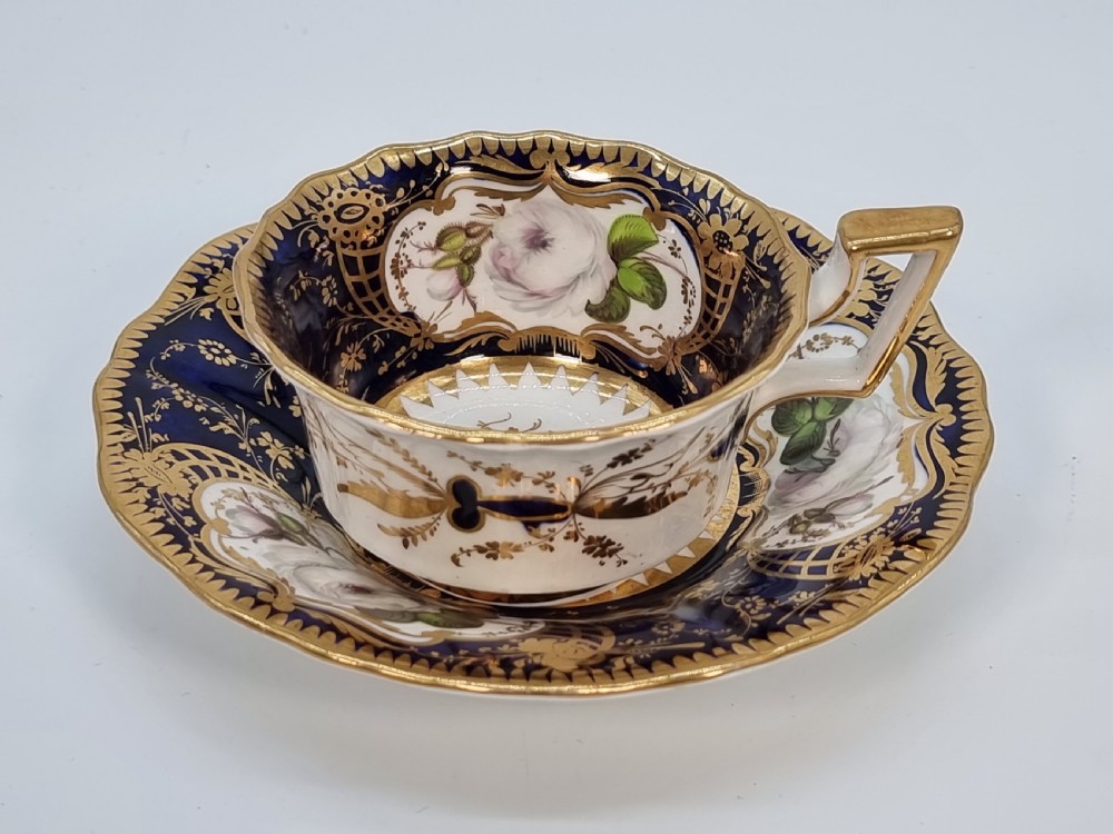 An early nineteenth-century Coalport porcelain cabinet trio featuring a teacup, coffee cup, and saucer styled with cobalt blue borders, gilded detailing, and hand-painted white rose panels. View XXVIII