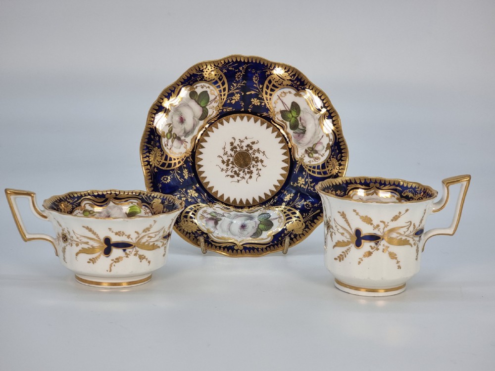An early nineteenth-century Coalport porcelain cabinet trio featuring a teacup, coffee cup, and saucer styled with cobalt blue borders, gilded detailing, and hand-painted white rose panels. View XXX