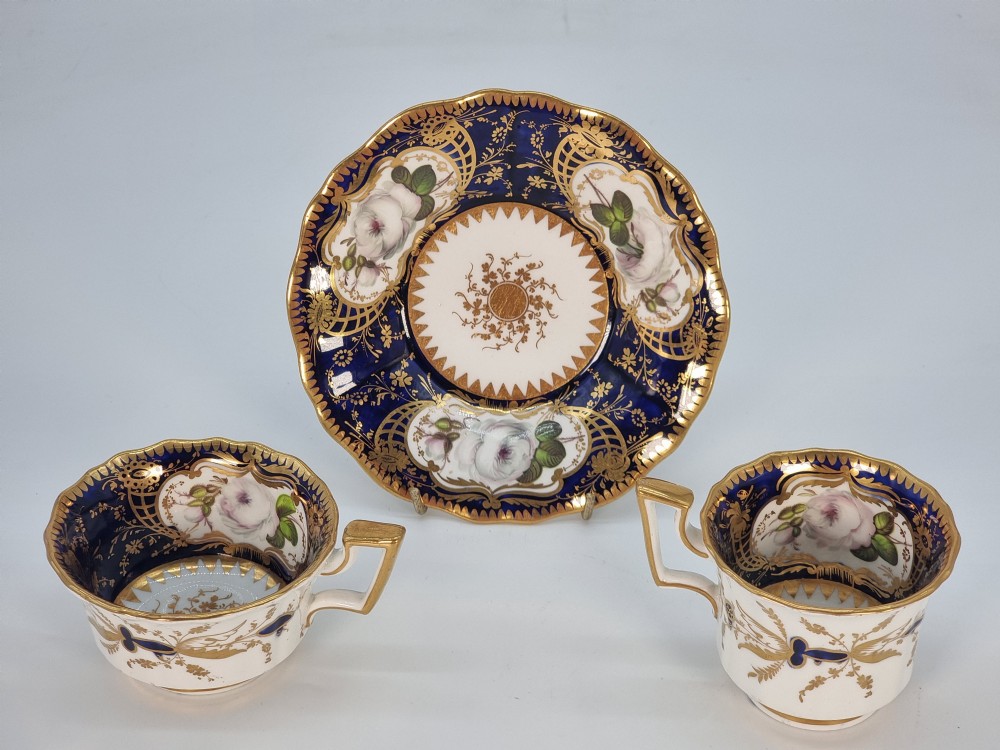 An early nineteenth-century Coalport porcelain cabinet trio featuring a teacup, coffee cup, and saucer styled with cobalt blue borders, gilded detailing, and hand-painted white rose panels.