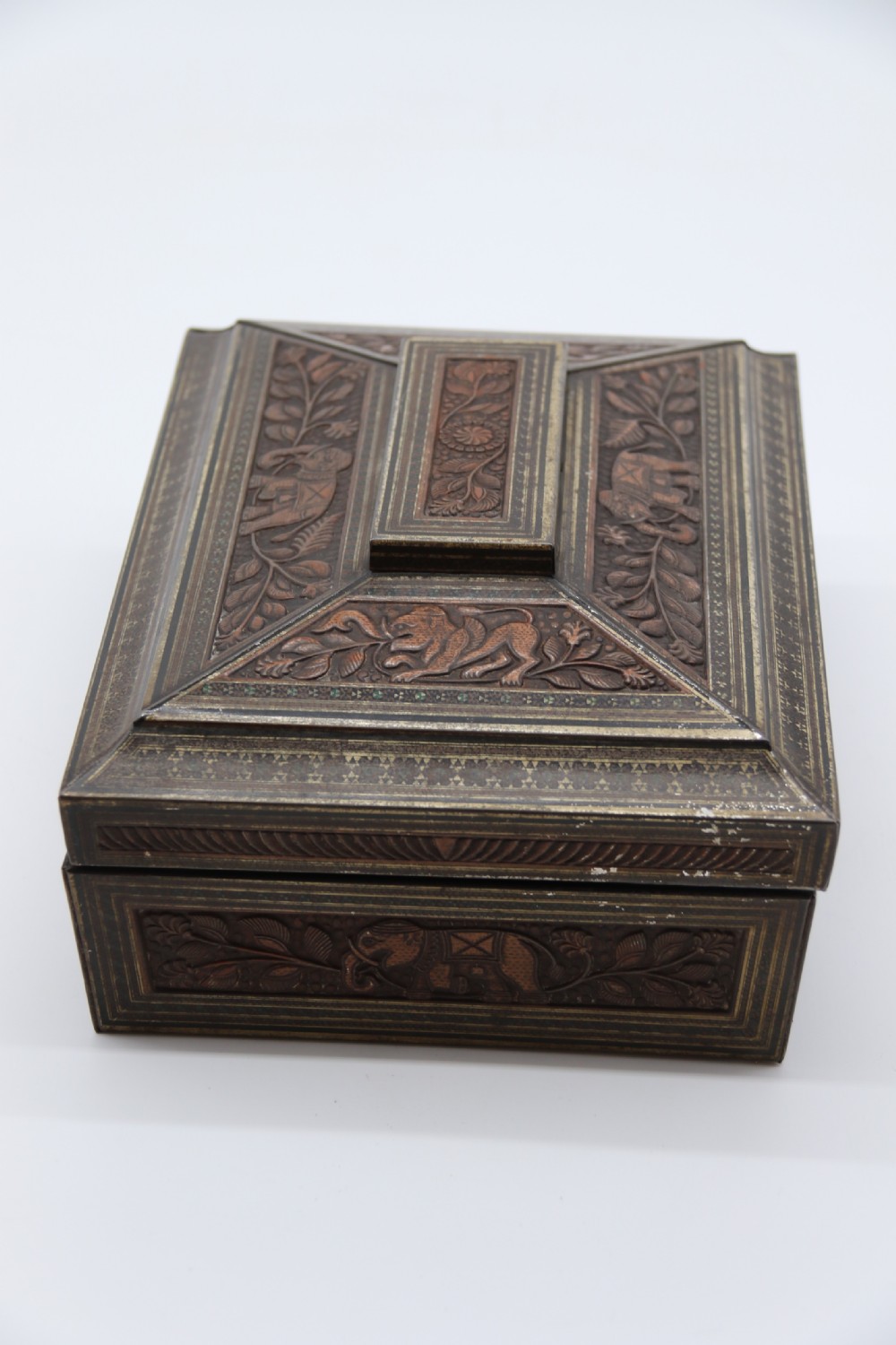 A rectangular lithographed tinplate box with a stepped lid, featuring embossed floral and elephant motifs designed to simulate carved wood. View IV