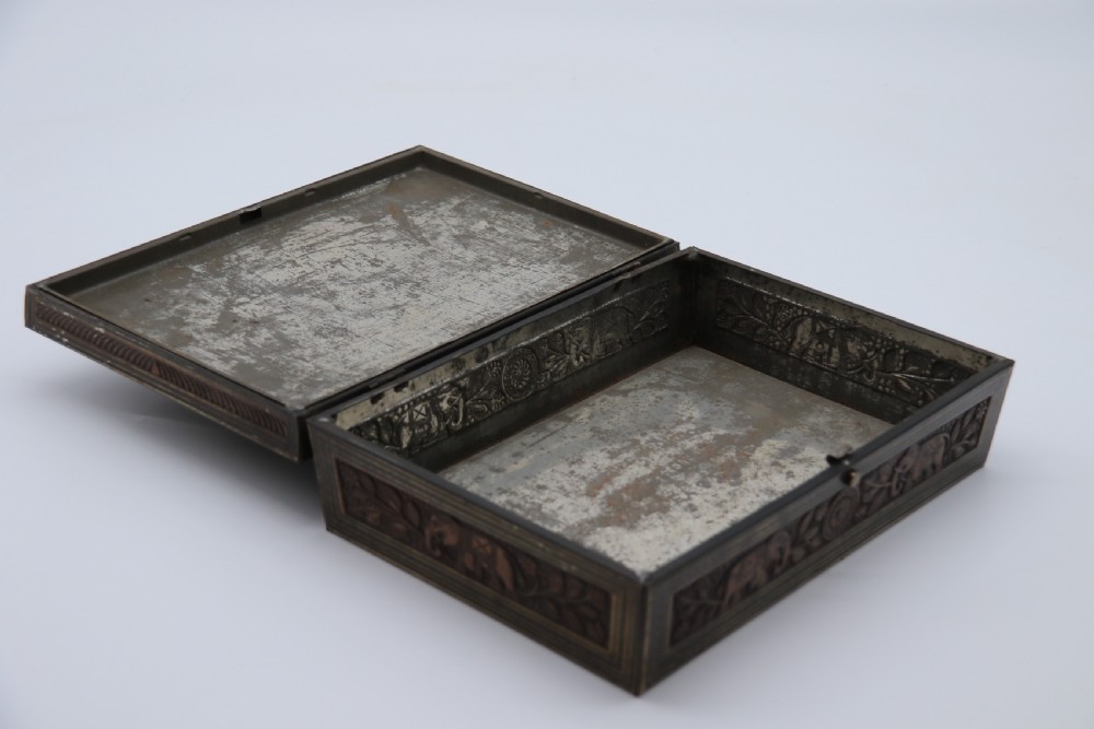 A rectangular lithographed tinplate box with a stepped lid, featuring embossed floral and elephant motifs designed to simulate carved wood. View X