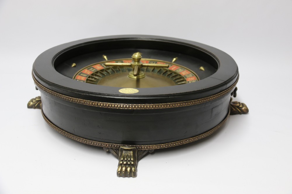 An antique French roulette wheel with ebonised wood casing and brass claw feet, set in front of an original dark green felt layout cloth printed with gold French gaming text. View VIII