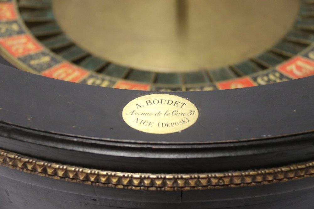 An antique French roulette wheel with ebonised wood casing and brass claw feet, set in front of an original dark green felt layout cloth printed with gold French gaming text. View XI
