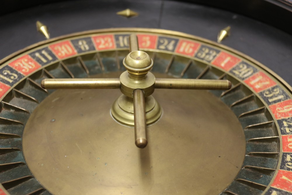 An antique French roulette wheel with ebonised wood casing and brass claw feet, set in front of an original dark green felt layout cloth printed with gold French gaming text. View XII