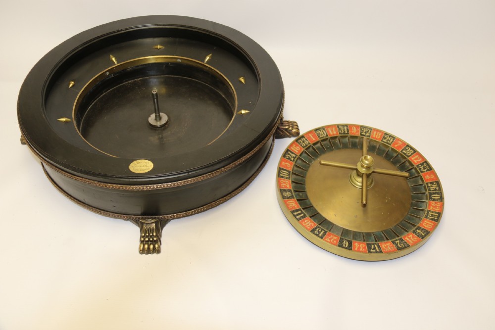 An antique French roulette wheel with ebonised wood casing and brass claw feet, set in front of an original dark green felt layout cloth printed with gold French gaming text. View XIV