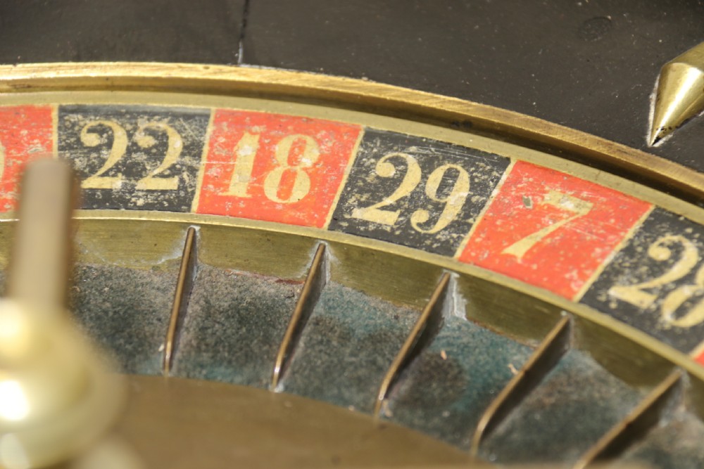 An antique French roulette wheel with ebonised wood casing and brass claw feet, set in front of an original dark green felt layout cloth printed with gold French gaming text. View XVII