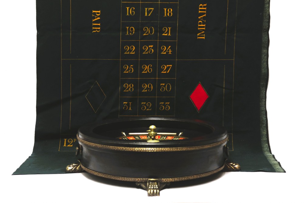 An antique French roulette wheel with ebonised wood casing and brass claw feet, set in front of an original dark green felt layout cloth printed with gold French gaming text. View XX