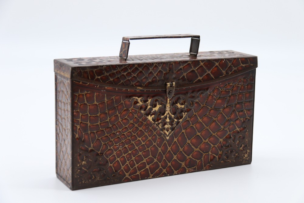A rectangular metal novelty biscuit tin shaped like a leather satchel with printed crocodile texture, gold scroll accents, and a top carrying handle.