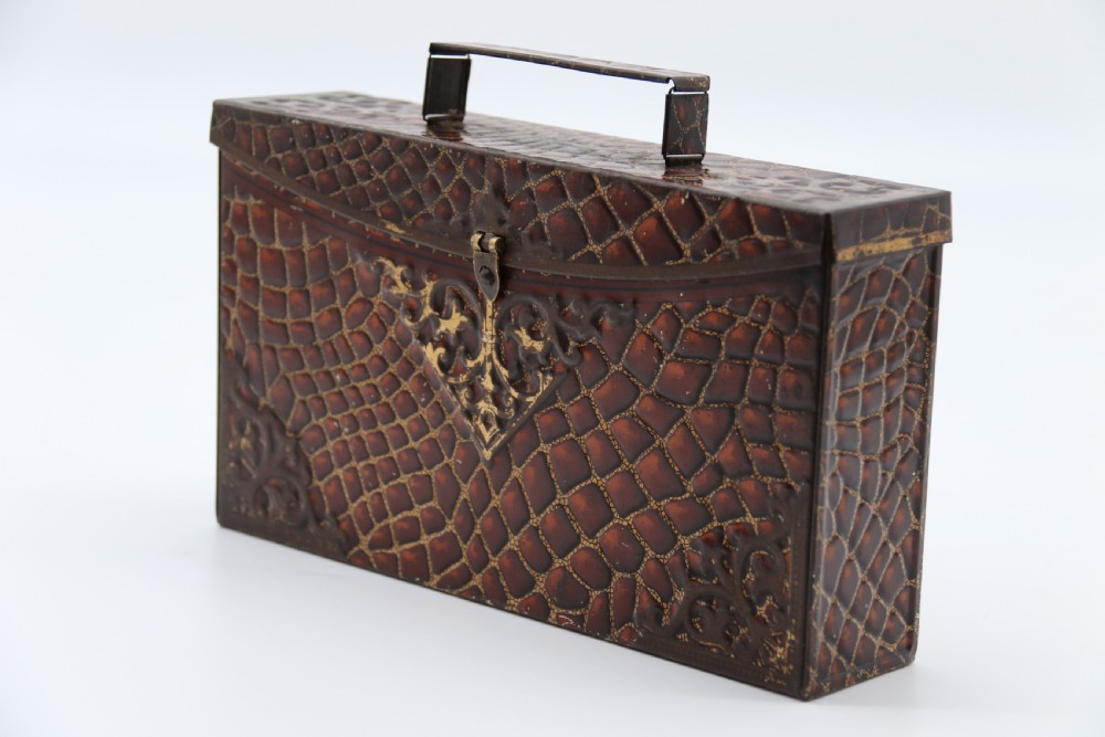 A rectangular metal novelty biscuit tin shaped like a leather satchel with printed crocodile texture, gold scroll accents, and a top carrying handle. View IV