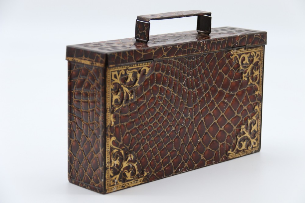 A rectangular metal novelty biscuit tin shaped like a leather satchel with printed crocodile texture, gold scroll accents, and a top carrying handle. View VI