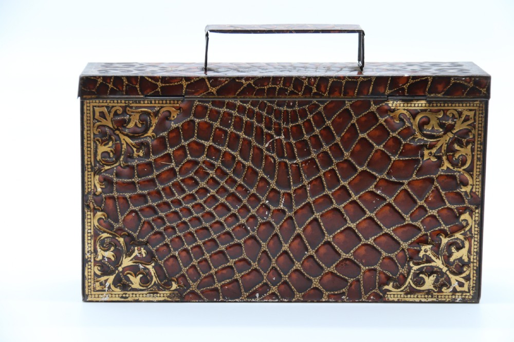 A rectangular metal novelty biscuit tin shaped like a leather satchel with printed crocodile texture, gold scroll accents, and a top carrying handle. View VII