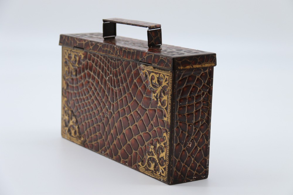 A rectangular metal novelty biscuit tin shaped like a leather satchel with printed crocodile texture, gold scroll accents, and a top carrying handle. View VIII