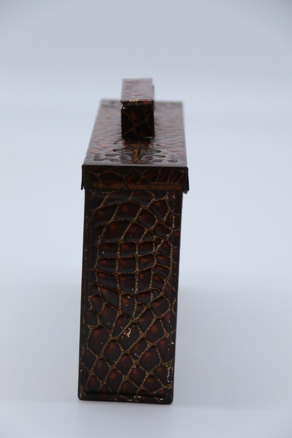 A rectangular metal novelty biscuit tin shaped like a leather satchel with printed crocodile texture, gold scroll accents, and a top carrying handle. View IX