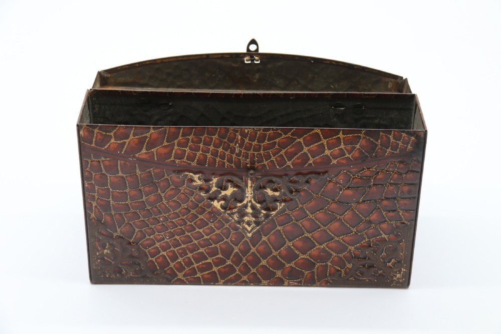 A rectangular metal novelty biscuit tin shaped like a leather satchel with printed crocodile texture, gold scroll accents, and a top carrying handle. View X