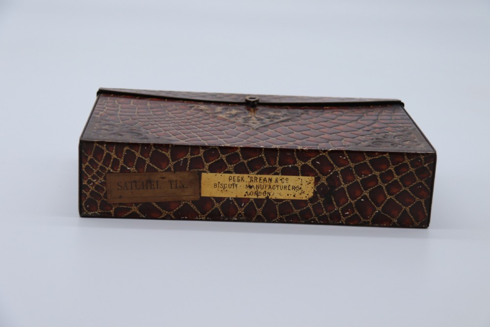 A rectangular metal novelty biscuit tin shaped like a leather satchel with printed crocodile texture, gold scroll accents, and a top carrying handle. View XII