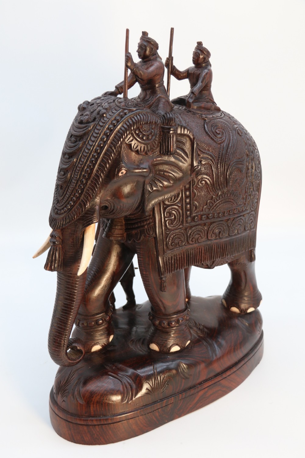 A dark hardwood sculpture depicting a ceremonial elephant adorned with decorative drapery accompanied by three mahouts on an integrated wooden base. View III