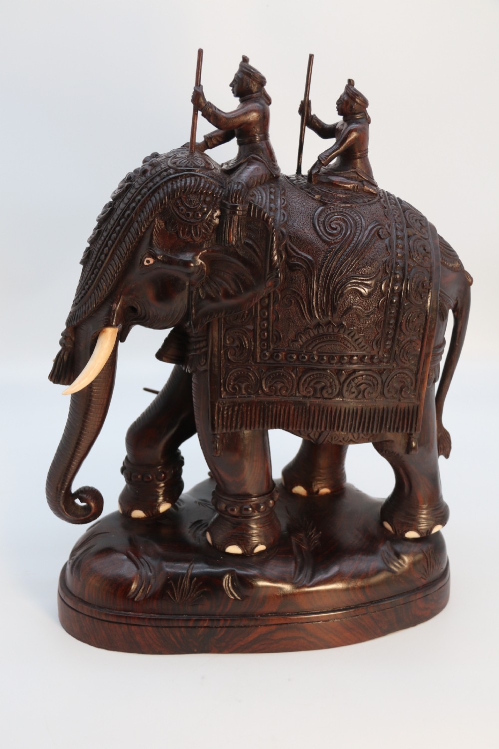 A dark hardwood sculpture depicting a ceremonial elephant adorned with decorative drapery accompanied by three mahouts on an integrated wooden base. View IV