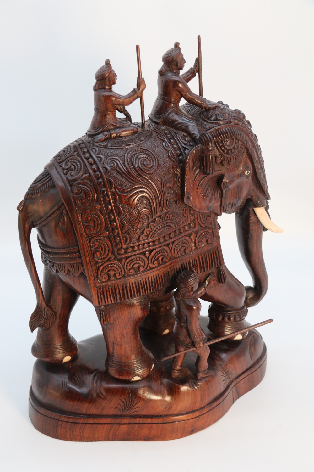 A dark hardwood sculpture depicting a ceremonial elephant adorned with decorative drapery accompanied by three mahouts on an integrated wooden base. View IX