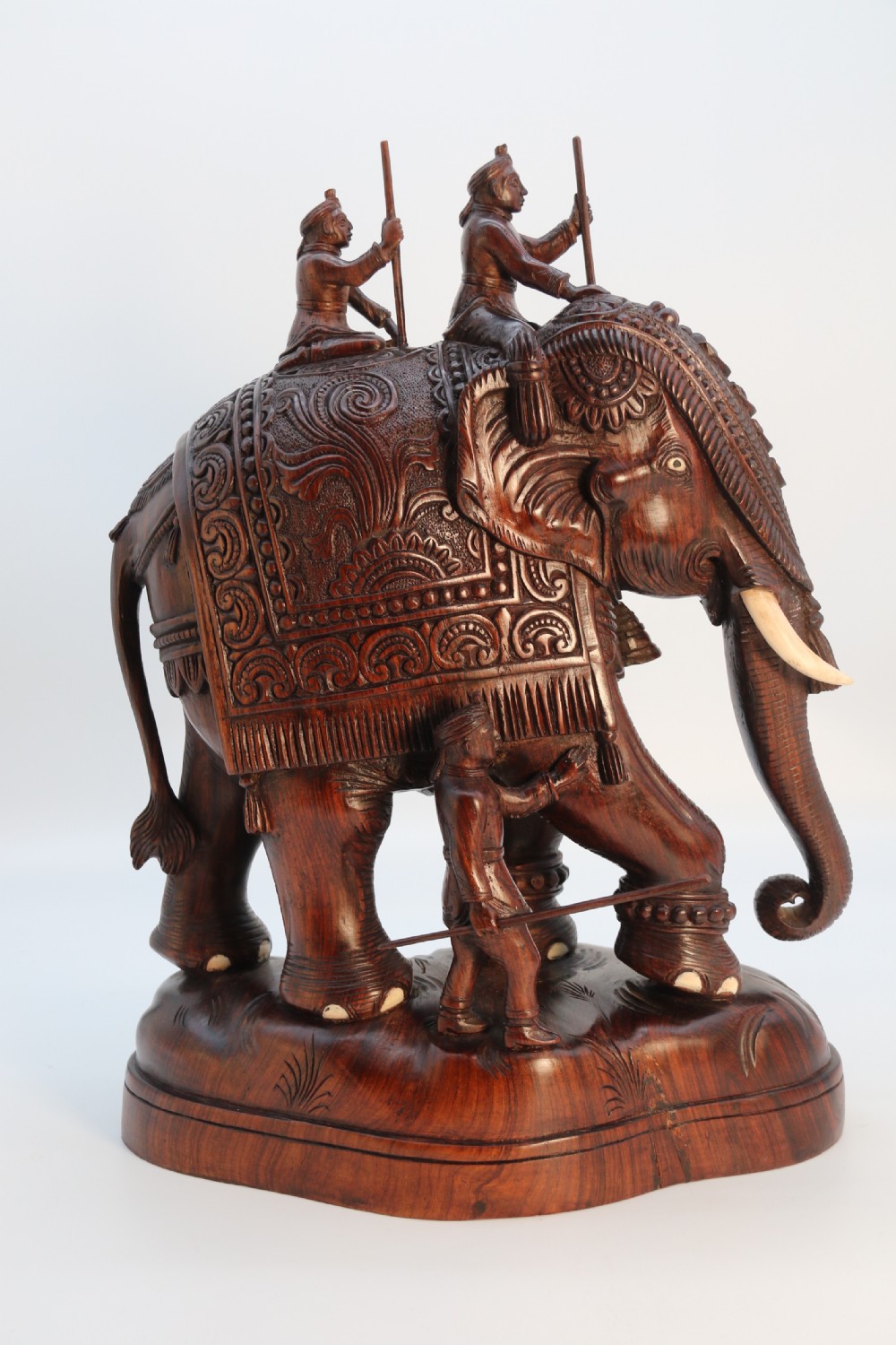A dark hardwood sculpture depicting a ceremonial elephant adorned with decorative drapery accompanied by three mahouts on an integrated wooden base. View X