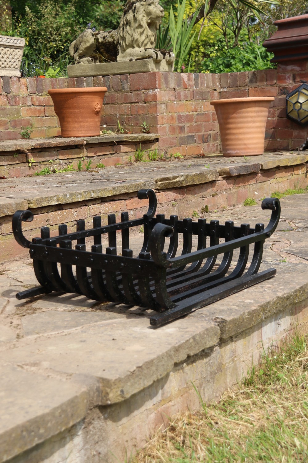 A black wrought iron fire basket featuring a curved slatted log cradle, scrolled corner terminals, and riveted front bars. View XVII
