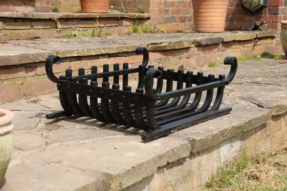 A black wrought iron fire basket featuring a curved slatted log cradle, scrolled corner terminals, and riveted front bars. View XVIII