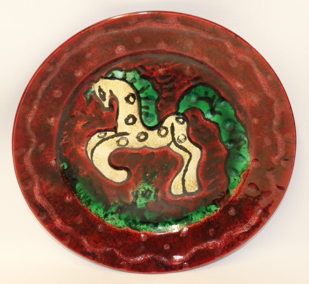 A circular glazed ceramic charger featuring a stylised cream horse motif against a deep red and emerald green reactive glaze.