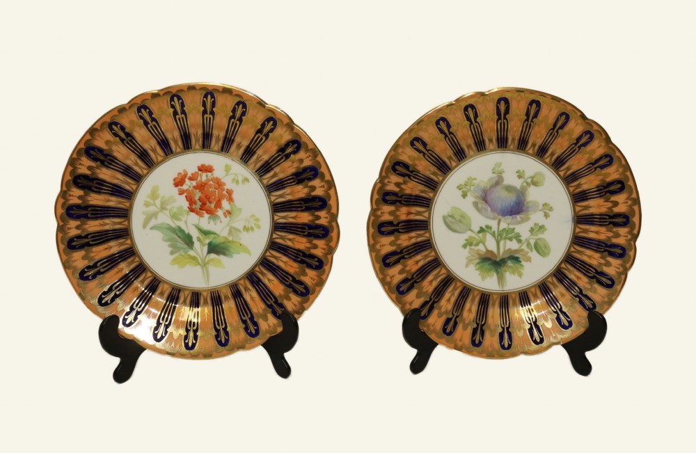 A pair of circular antique porcelain cabinet plates featuring central hand-painted floral motifs surrounded by elaborate gilded and cobalt blue borders.