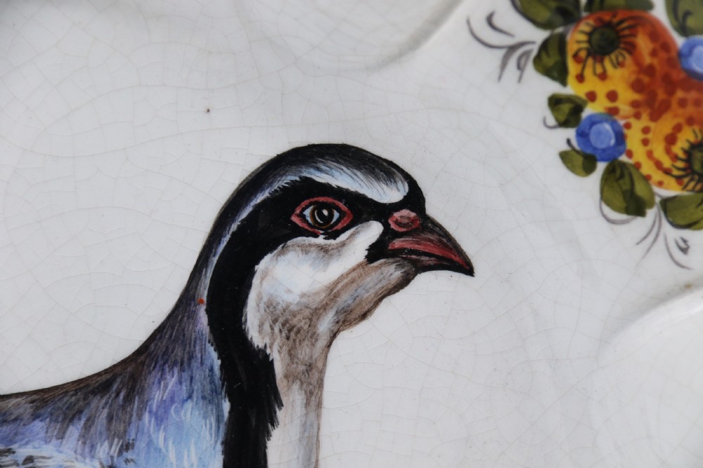 A circular tin-glazed earthenware wall charger featuring a hand-painted game bird in the centre surrounded by a scalloped, floral-decorated rim with blue edging. View X