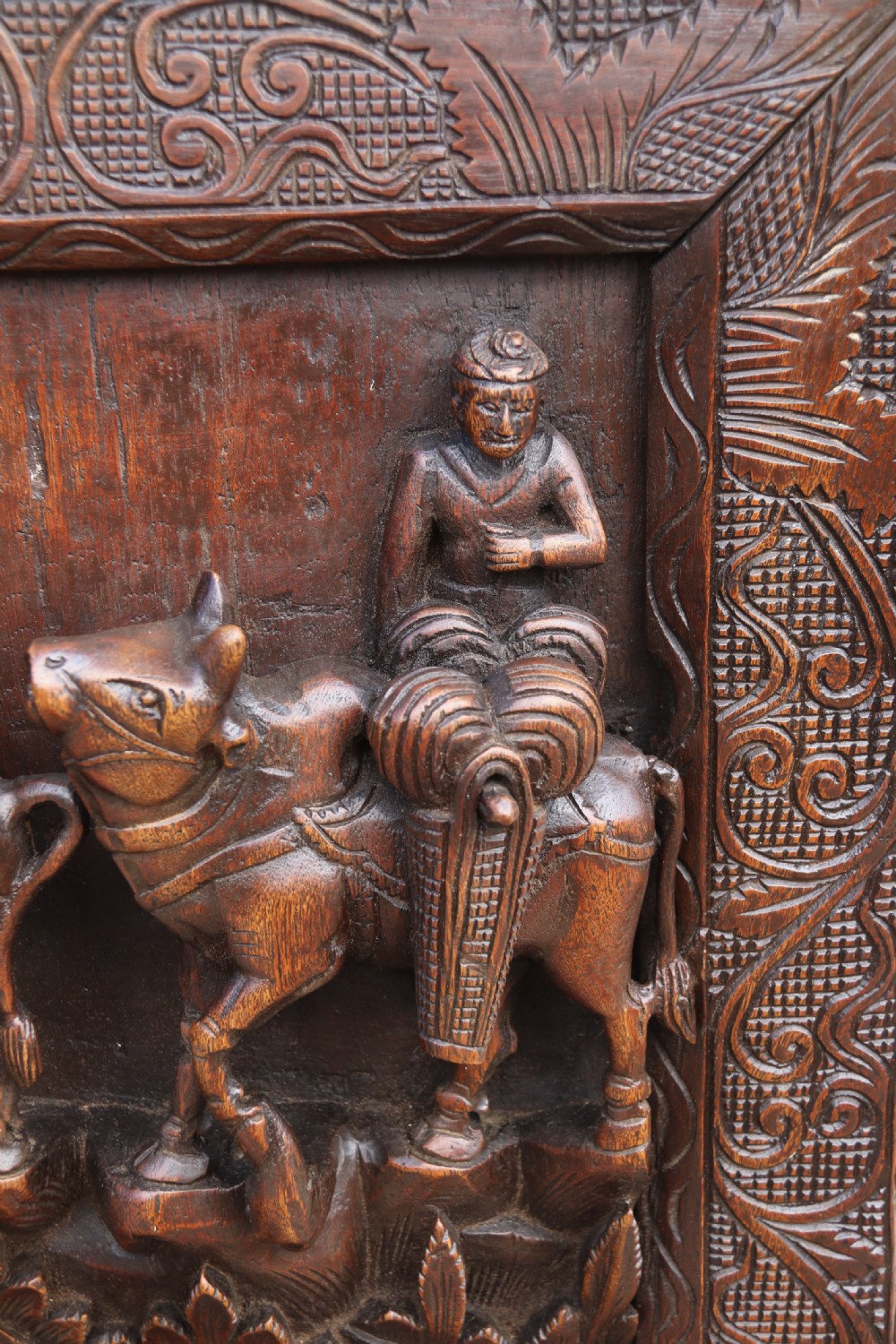 A dark hardwood rectangular panel featuring high-relief carvings of figures riding bullocks and a cart within an ornate carved border. View II