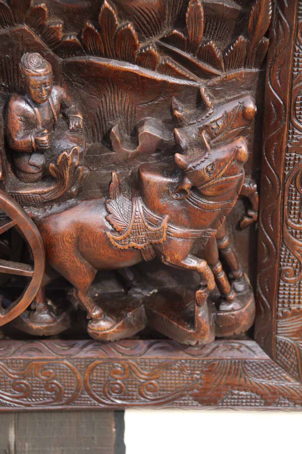 A dark hardwood rectangular panel featuring high-relief carvings of figures riding bullocks and a cart within an ornate carved border. View III