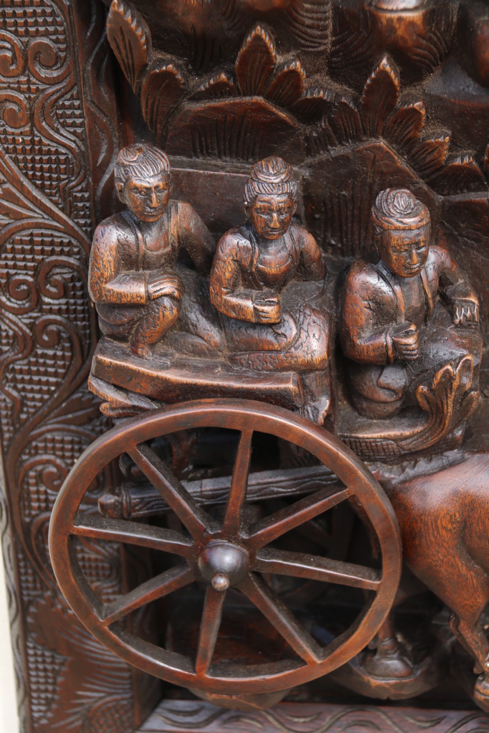 A dark hardwood rectangular panel featuring high-relief carvings of figures riding bullocks and a cart within an ornate carved border. View IV