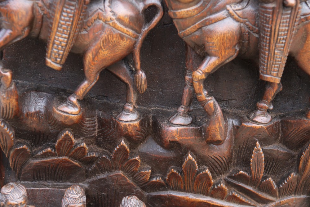 A dark hardwood rectangular panel featuring high-relief carvings of figures riding bullocks and a cart within an ornate carved border. View VI