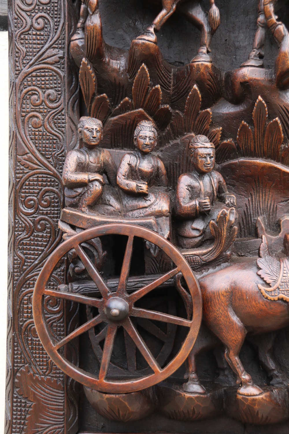 A dark hardwood rectangular panel featuring high-relief carvings of figures riding bullocks and a cart within an ornate carved border. View VIII
