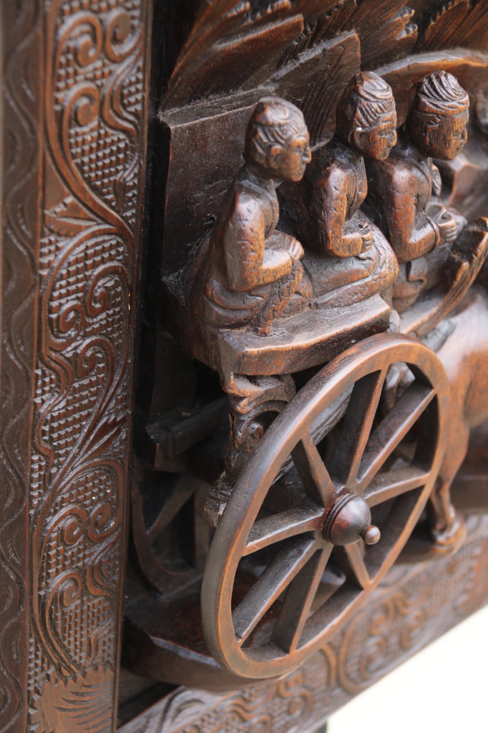 A dark hardwood rectangular panel featuring high-relief carvings of figures riding bullocks and a cart within an ornate carved border. View X