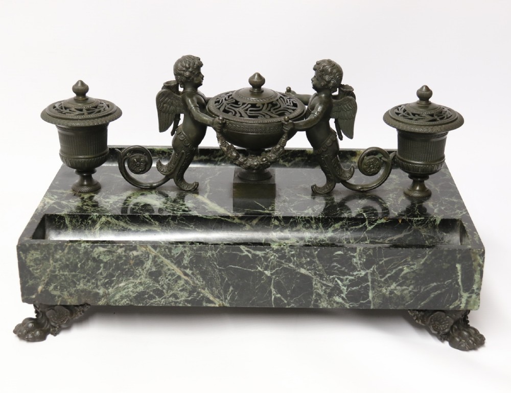 An Empire period dark green marble inkstand featuring dark patinated bronze cherub figures, twin urn inkwells, and carved claw feet.