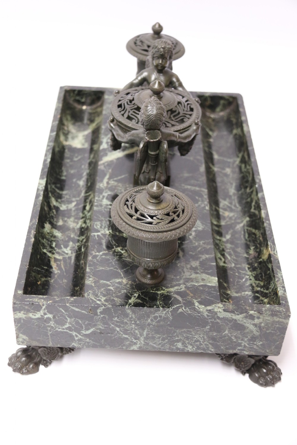 An Empire period dark green marble inkstand featuring dark patinated bronze cherub figures, twin urn inkwells, and carved claw feet. View X