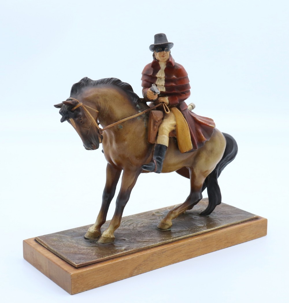 An Art Deco cold-painted metal figure of a masked highwayman on horseback mounted on a rectangular wooden plinth, featuring an integrated table lighter mechanism.